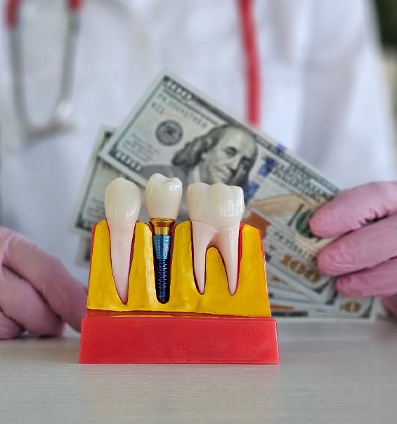 Gloved hands holding $100 bills next to model dental implant
